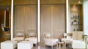 A hotel room with a large wooden wardrobe, a seating area with a sofa and chairs, a small table with a vase of flowers, and a shelf with bottles. - CA Hotel and Residence Phuket (Ratsada)