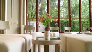 Habitación de hotel con dos camas, una mesa redonda con un jarrón de flores y ventanas amplias que dan a vistas de vegetación.
