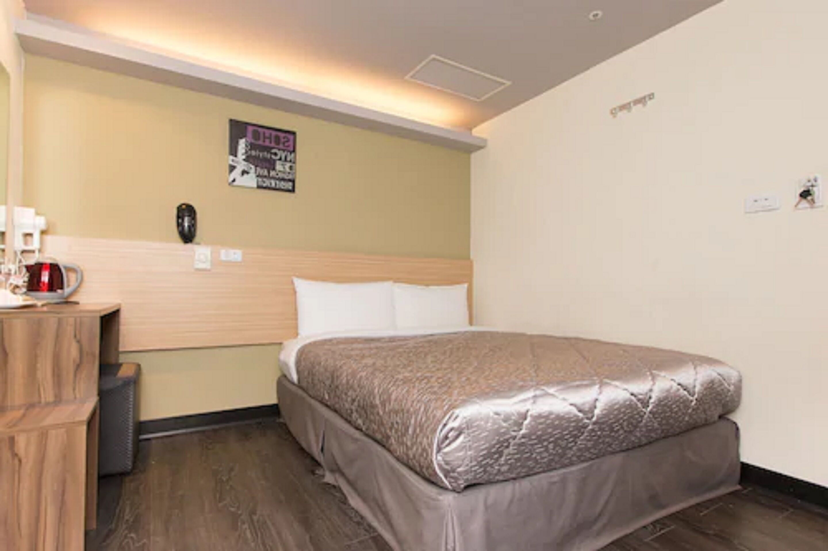 A hotel room with a bed, a wooden headboard, a nightstand with a red object, and a poster on the wall.