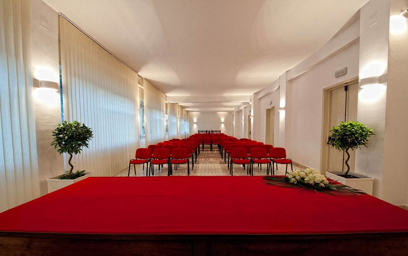 Un salón de conferencias con una larga mesa y sillas rojas, flanqueado por paredes blancas y plantas en macetas.