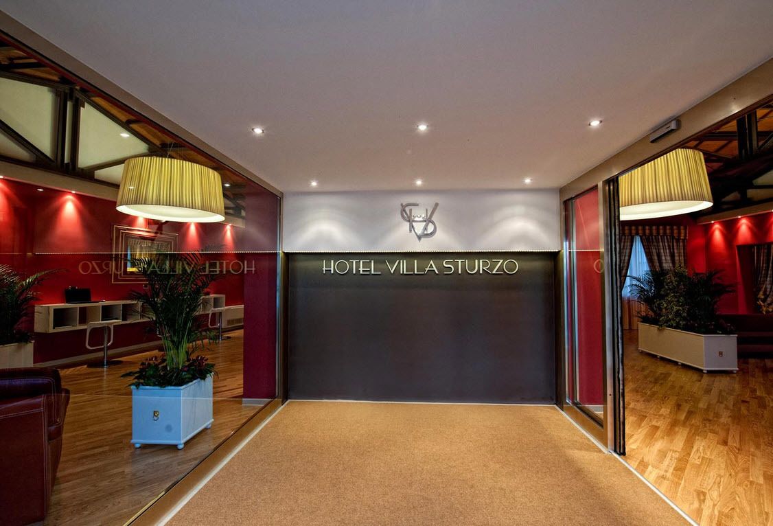 A modern hotel lobby with a reception desk, a large chandelier, and a sign reading "HOTEL VILLA STORZO".