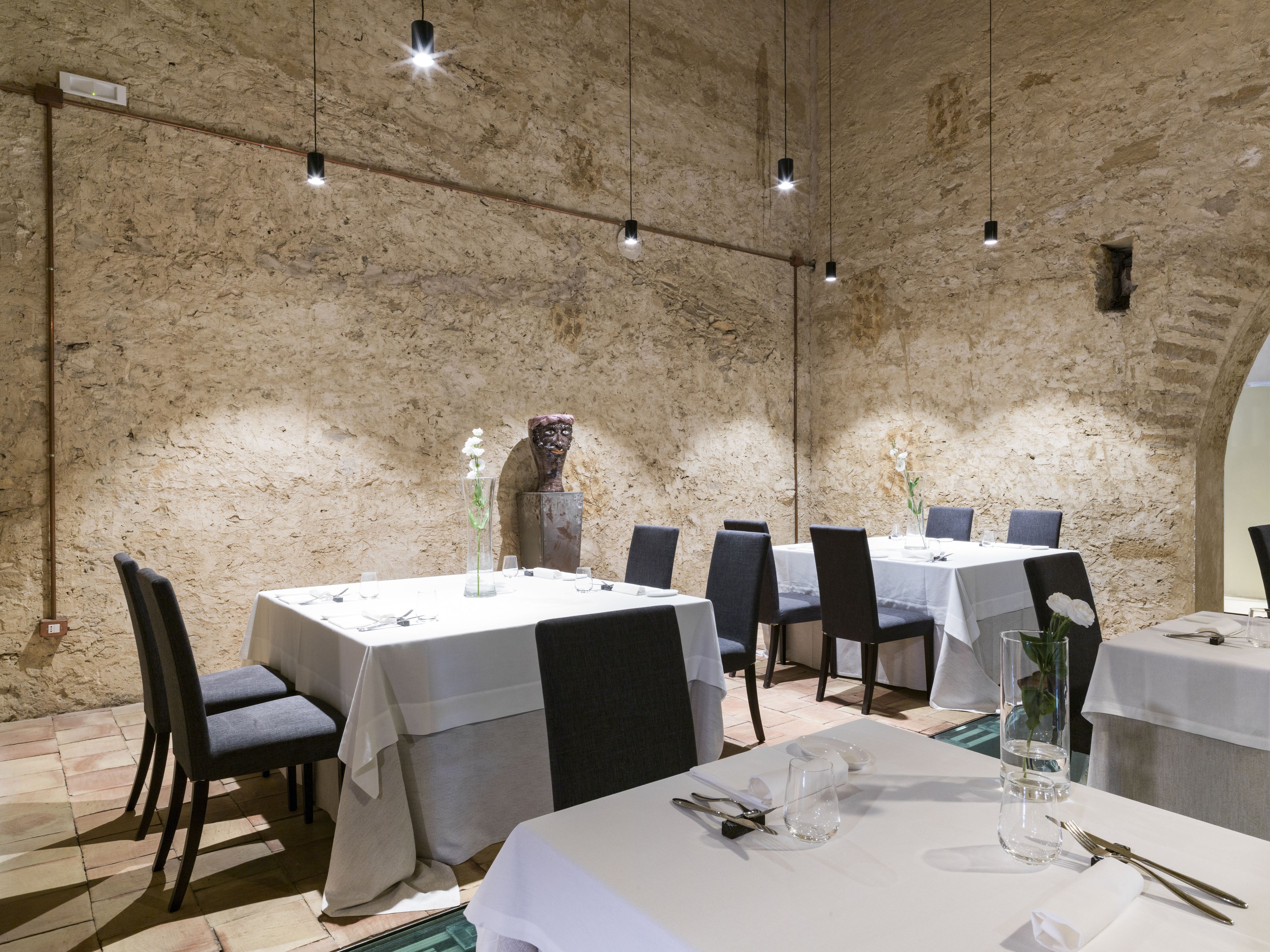 Ein Restaurant mit rustikalen Steinwänden, mehreren Tischen mit weißen Tischdecken und moderner Pendelleuchten.
