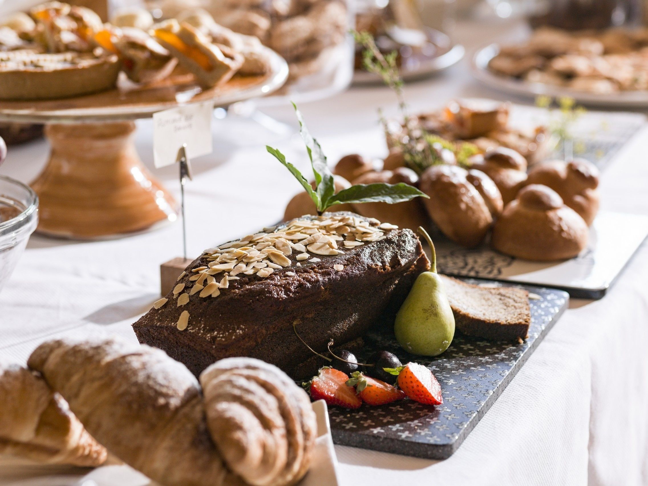 A variety of baked goods including a chocolate cake with almonds, pastries, and fresh fruits on a table.