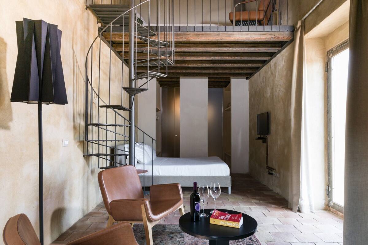 A room with a spiral staircase, a bed, a table with a book and glasses, and two chairs.