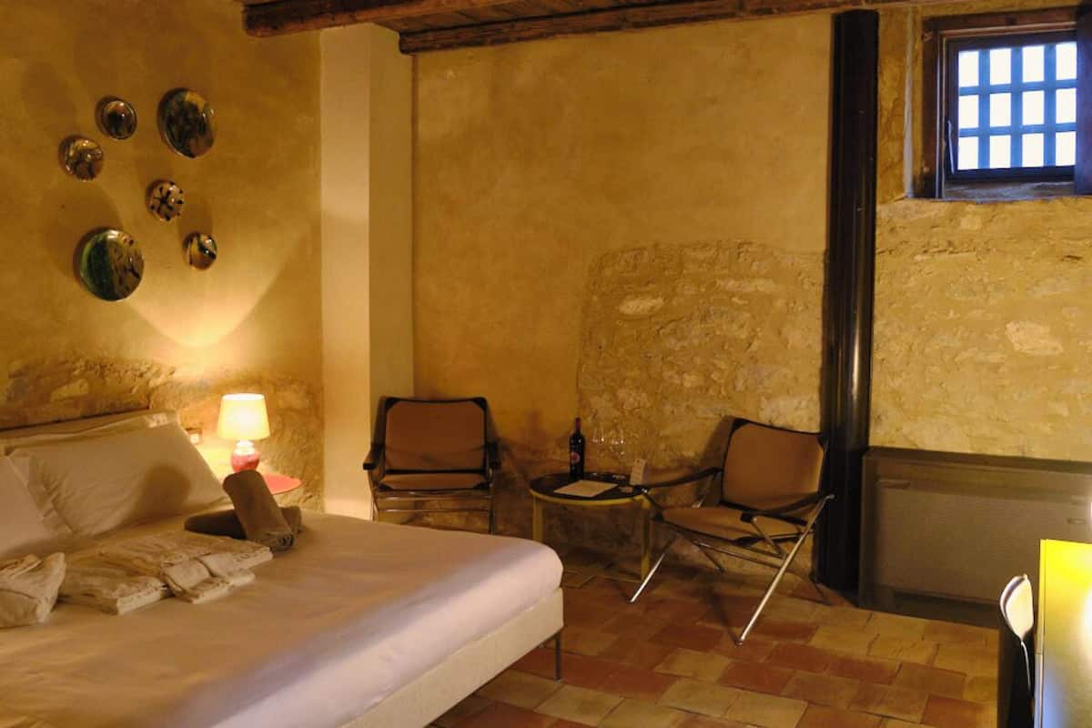 A cozy room with a bed, two chairs, a small table with a lamp, and a window.