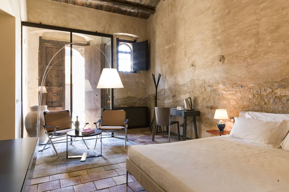 A rustic room with a bed, a desk, two chairs, a lamp, and a large vase.
