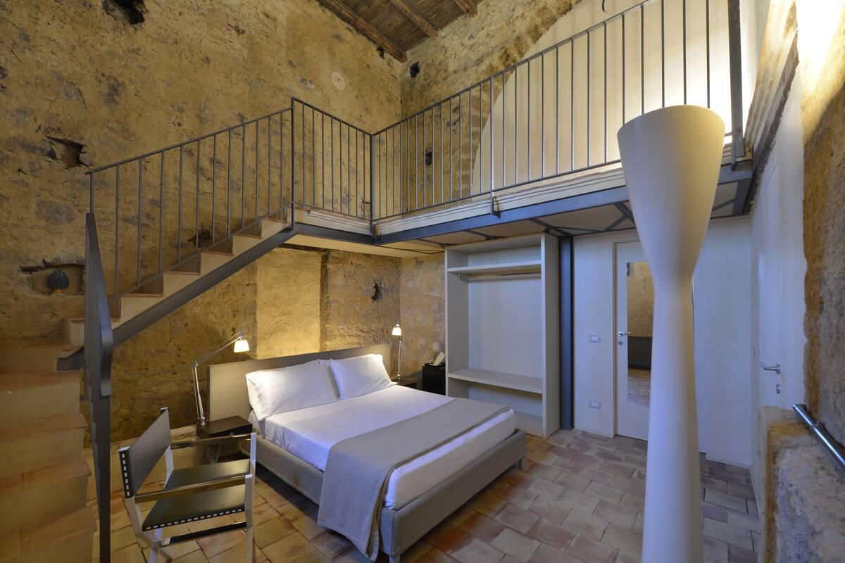 A bedroom with a bed, a chair, a desk, a lamp, a wardrobe, and a staircase.