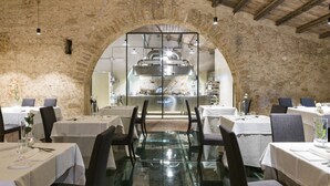 Un restaurant amb una paret de pedra rústica i una porta d'arc que dona a la cuina. Les taules estan preparades amb teles blanques i cadires.