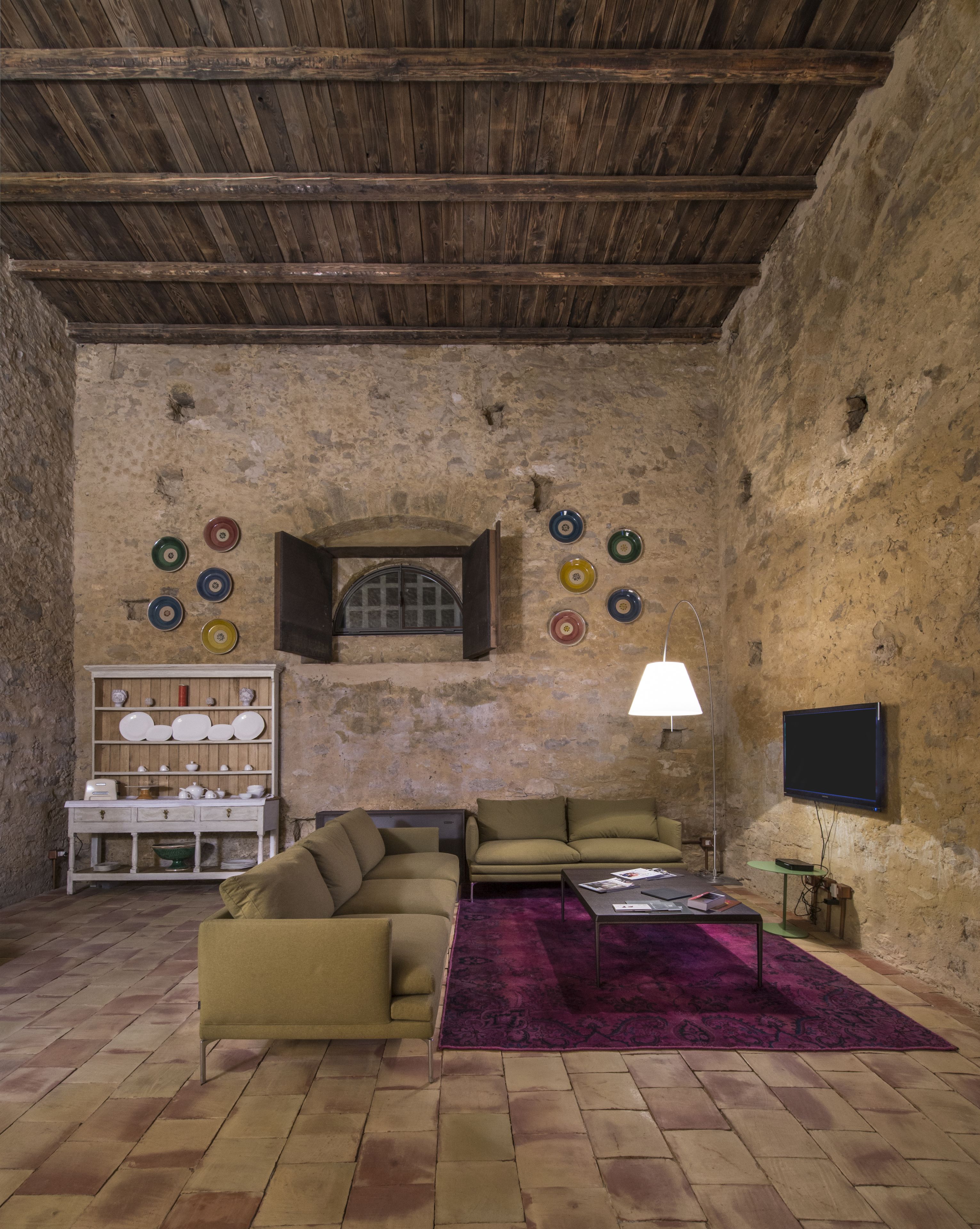 A rustic living room with a wooden ceiling, a sofa, a coffee table, a TV, and a shelf with decorative plates.