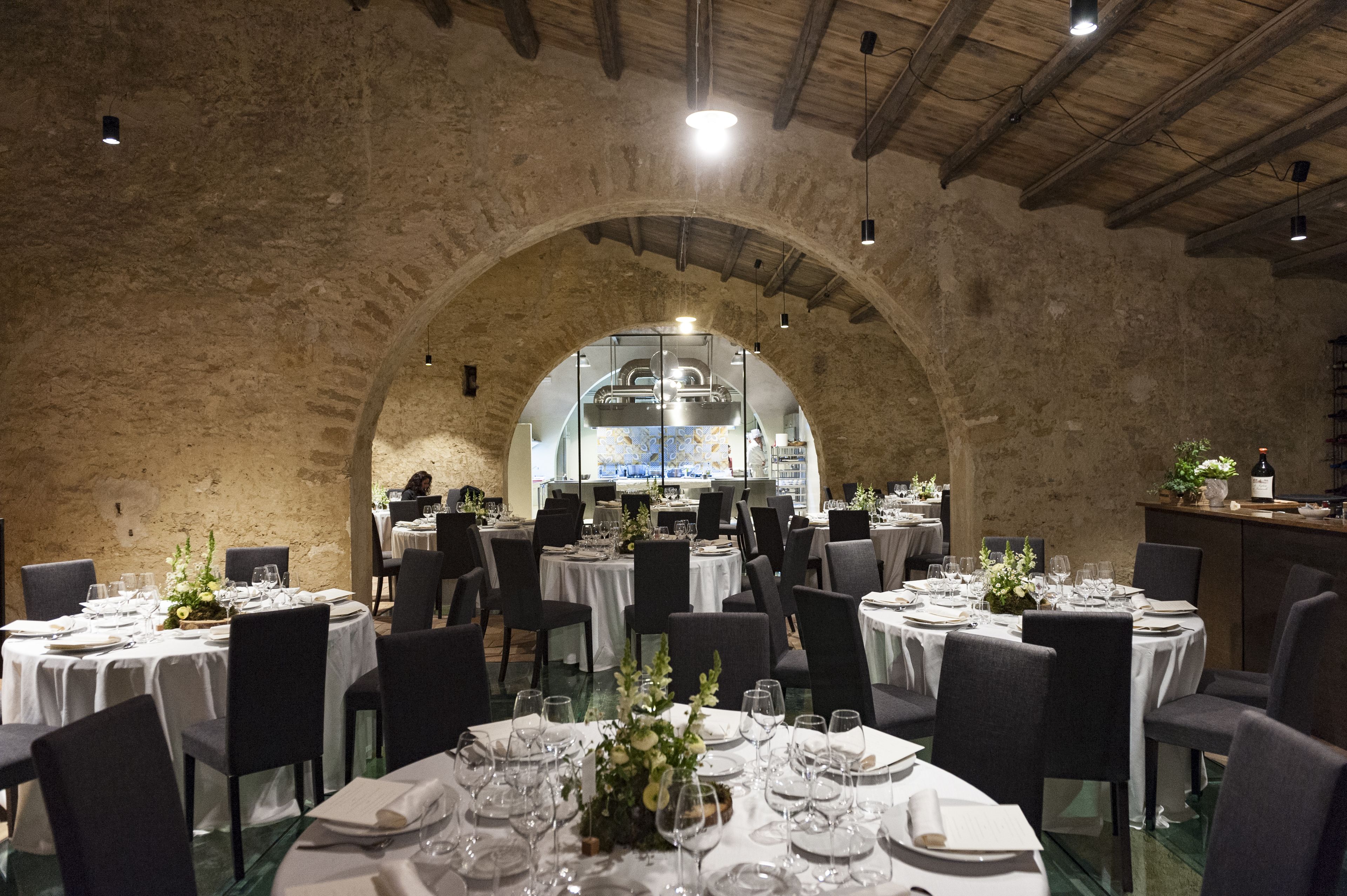 A restaurant with round tables set for dining, an arched stone ceiling, and a kitchen visible through the arch.