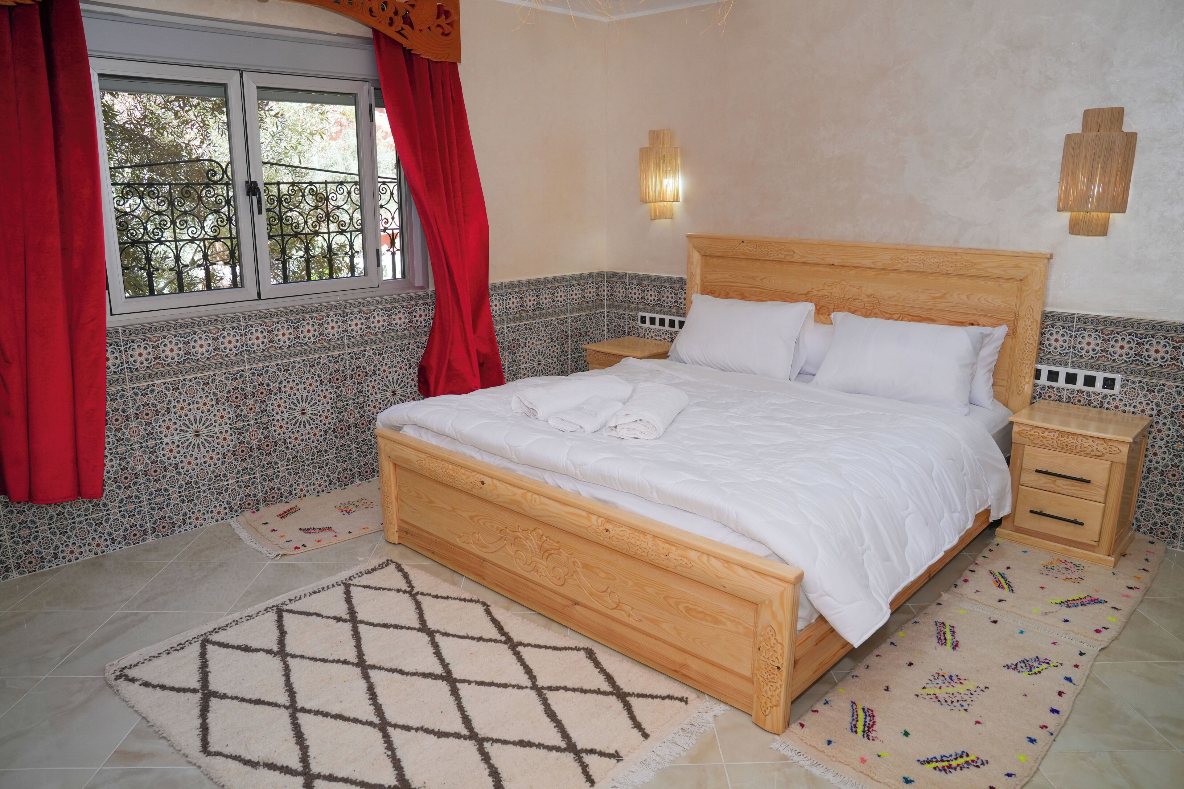 Un dormitorio con cama de madera, dos mesitas de noche, una ventana con cortinas rojas y paredes con baldosas estampadas.