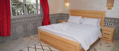 Un dormitorio con cama de madera, dos mesitas de noche, una ventana con cortinas rojas y paredes con baldosas estampadas.
