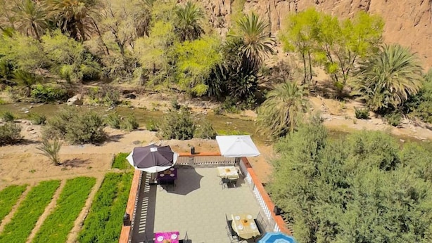 An outdoor patio area with tables and umbrellas, surrounded by greenery and a rocky cliff. - Auberge La Vallée (Toudgha El Oulia)