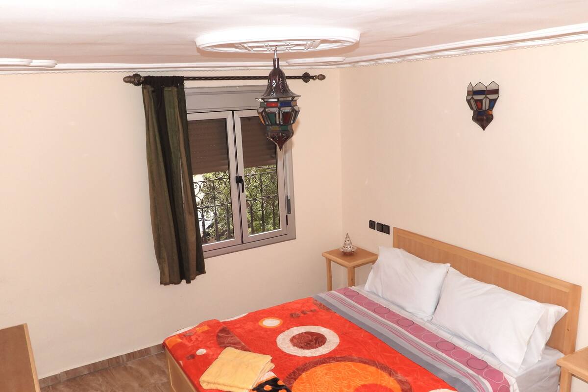 Een slaapkamer met een bed, een raam met gordijnen, een nachtkastje, een wandlamp en een plafondlamp.