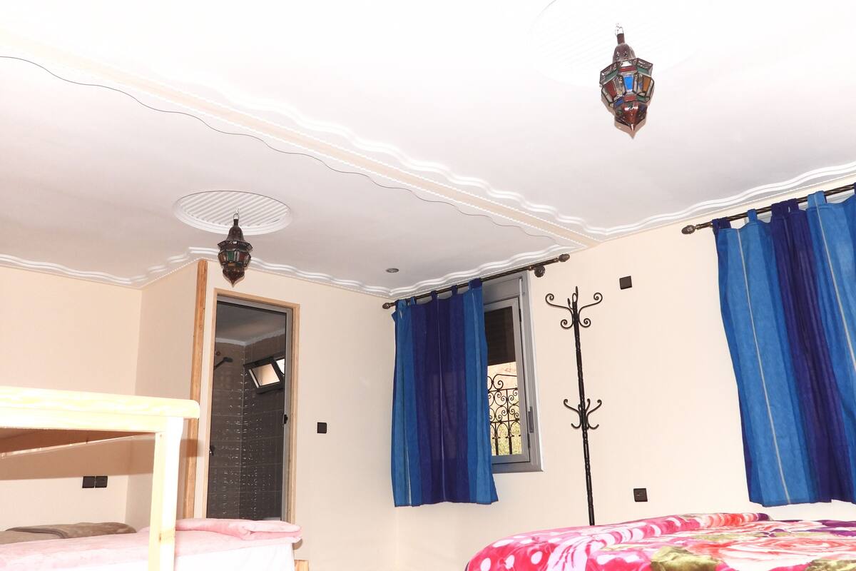 Een slaapkamer met een stapelbed, een raam met blauwe gordijnen, een kapstok aan de muur en een plafondlamp.