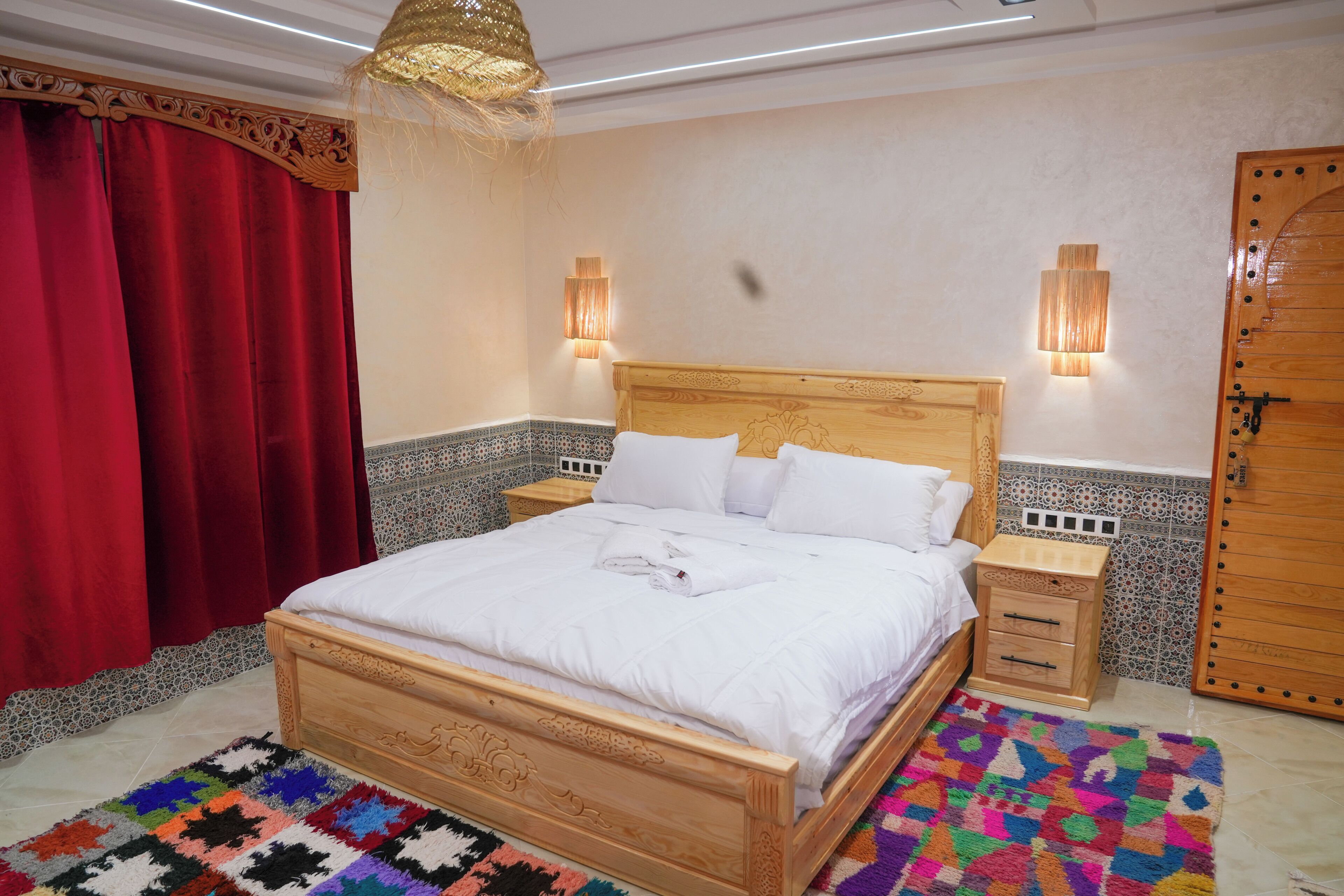 Una cama bien hecha con sábanas blancas, un cabecero de madera, dos mesitas de noche con lámparas, una alfombra colorida y una cortina roja.