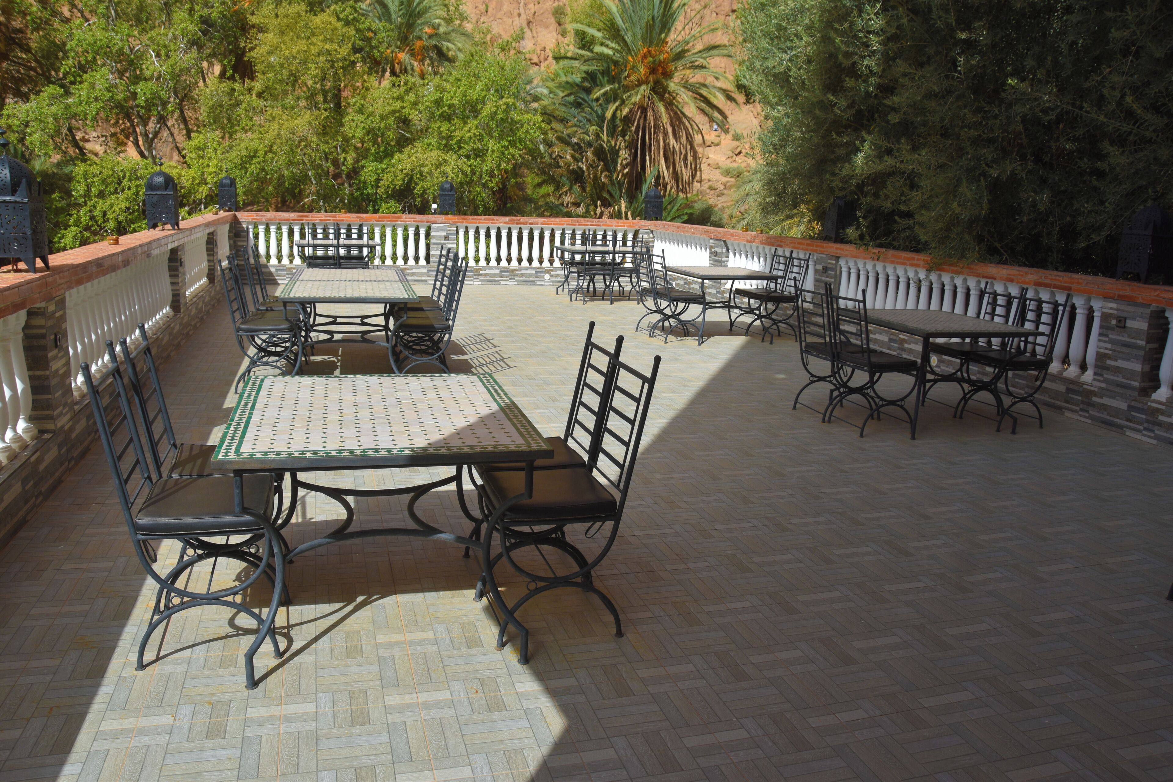 Eine Terrasse im Freien mit mehreren Tischen und Stühlen, umgeben von einer niedrigen Mauer und üppiger Bepflanzung.