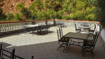 An outdoor patio with tables and chairs, surrounded by a white balustrade, set against a backdrop of green trees and a red rock hillside.