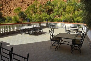 An outdoor patio with tables and chairs, surrounded by a white balustrade, set against a backdrop of green trees and a red rock hillside.