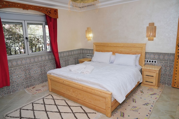 Quarto com cama de madeira, duas mesas de cabeceira, tapete estampado e vista para o exterior.