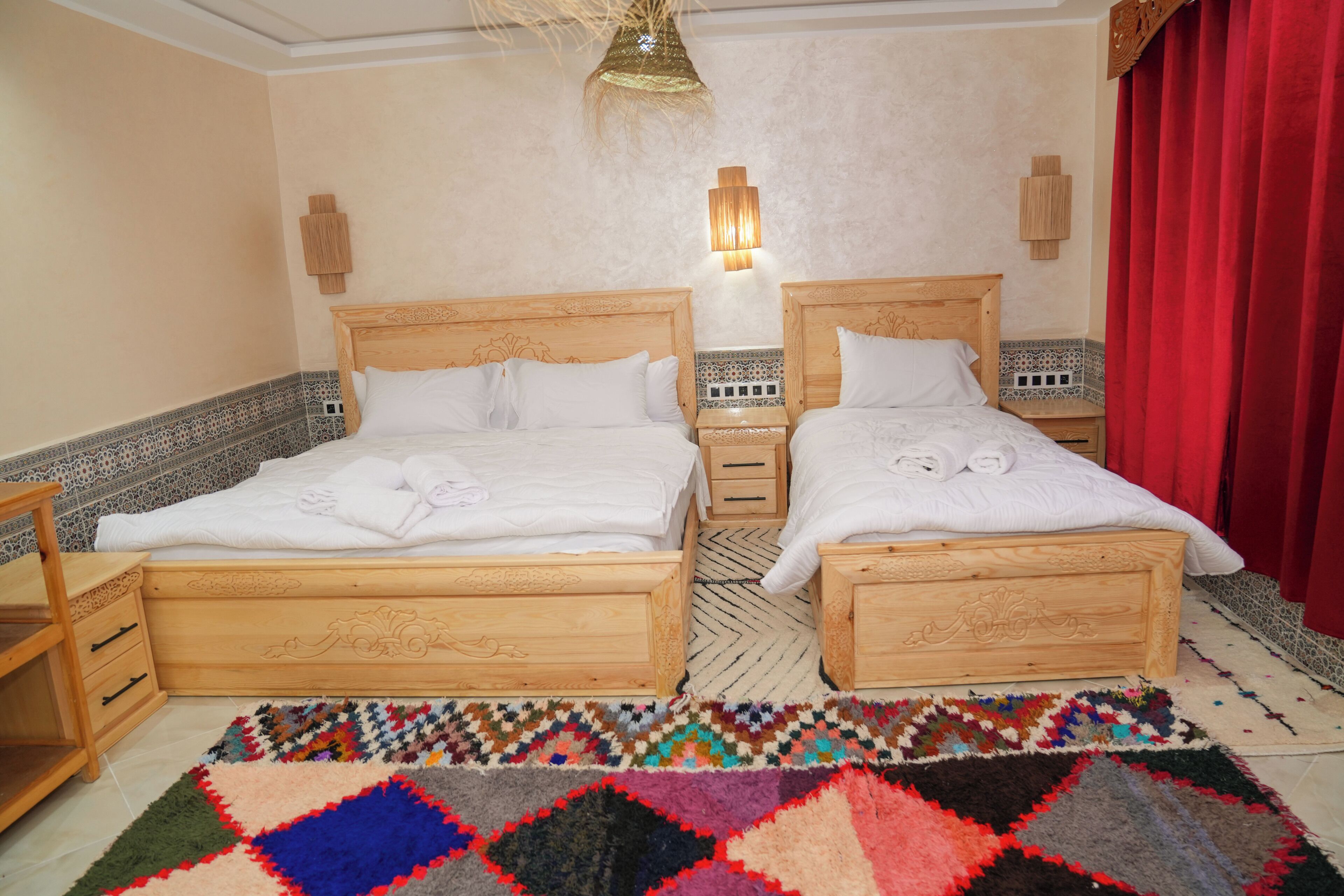 Quarto duplo com duas camas de madeira, um tapete estampado e luminária fixada na parede.