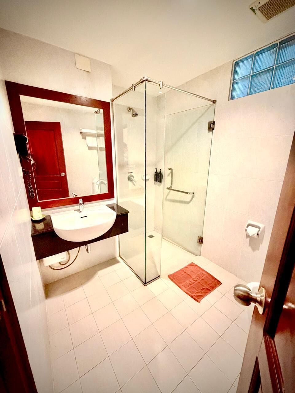 Un baño con cabina de ducha de vidrio, un lavamanos blanco, un espejo con marco rojo y un tapete rojo en el suelo.