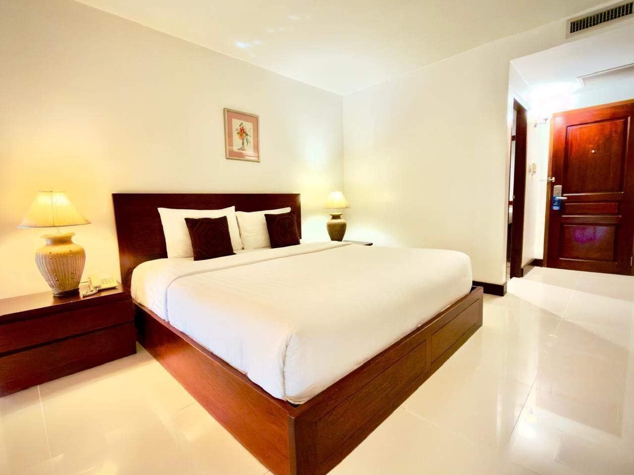 A hotel room with a large bed, two bedside tables with lamps, a wooden headboard, a framed picture on the wall, and a door to the right.