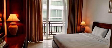 A hotel room with a bed, two bedside lamps, a wooden nightstand, a mirror, and a sliding glass door leading to a balcony.