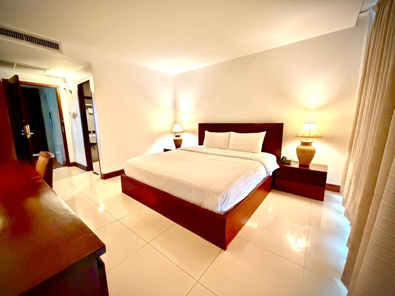 Una habitación de hotel con una cama grande, lámparas de noche, una mesita de noche de madera y una puerta que da a otra habitación.
