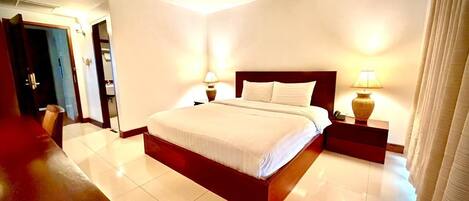 Una habitación de hotel con una cama grande, lámparas de noche, una mesita de noche de madera y una puerta que da a otra habitación.