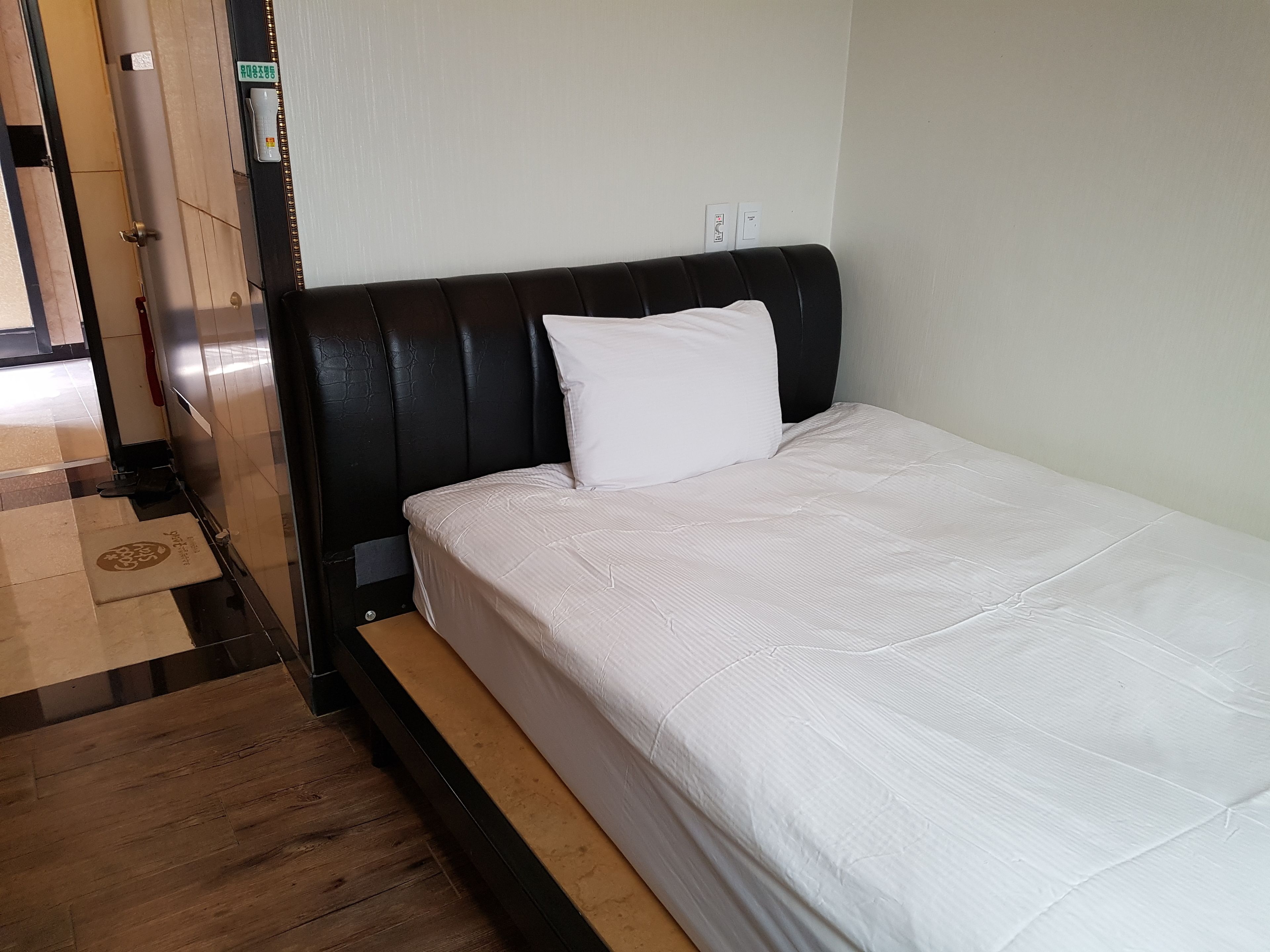 Uma cama bem arrumada com colchão e travesseiro brancos, cabeceira preta e piso de madeira.