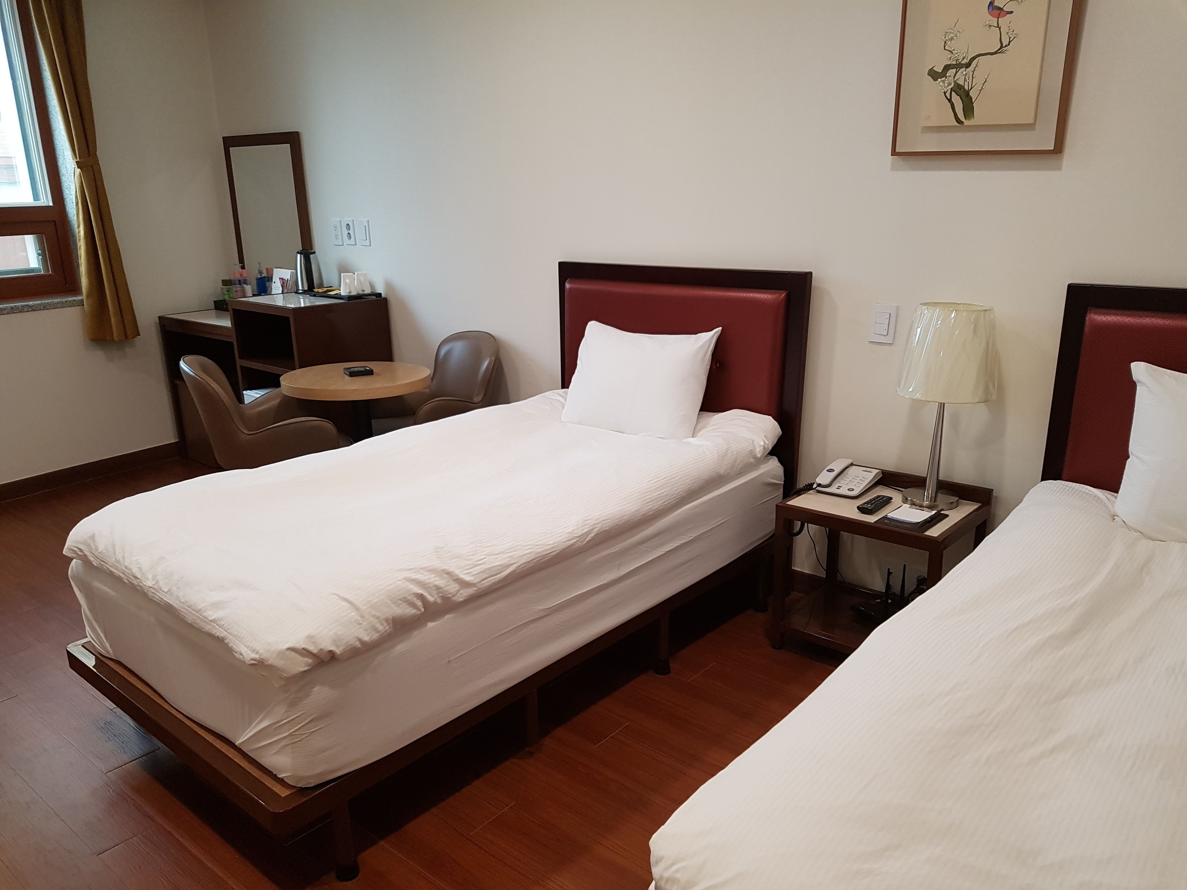 A hotel room with two single beds, a desk with a chair, a small table, a lamp, and a painting on the wall.
