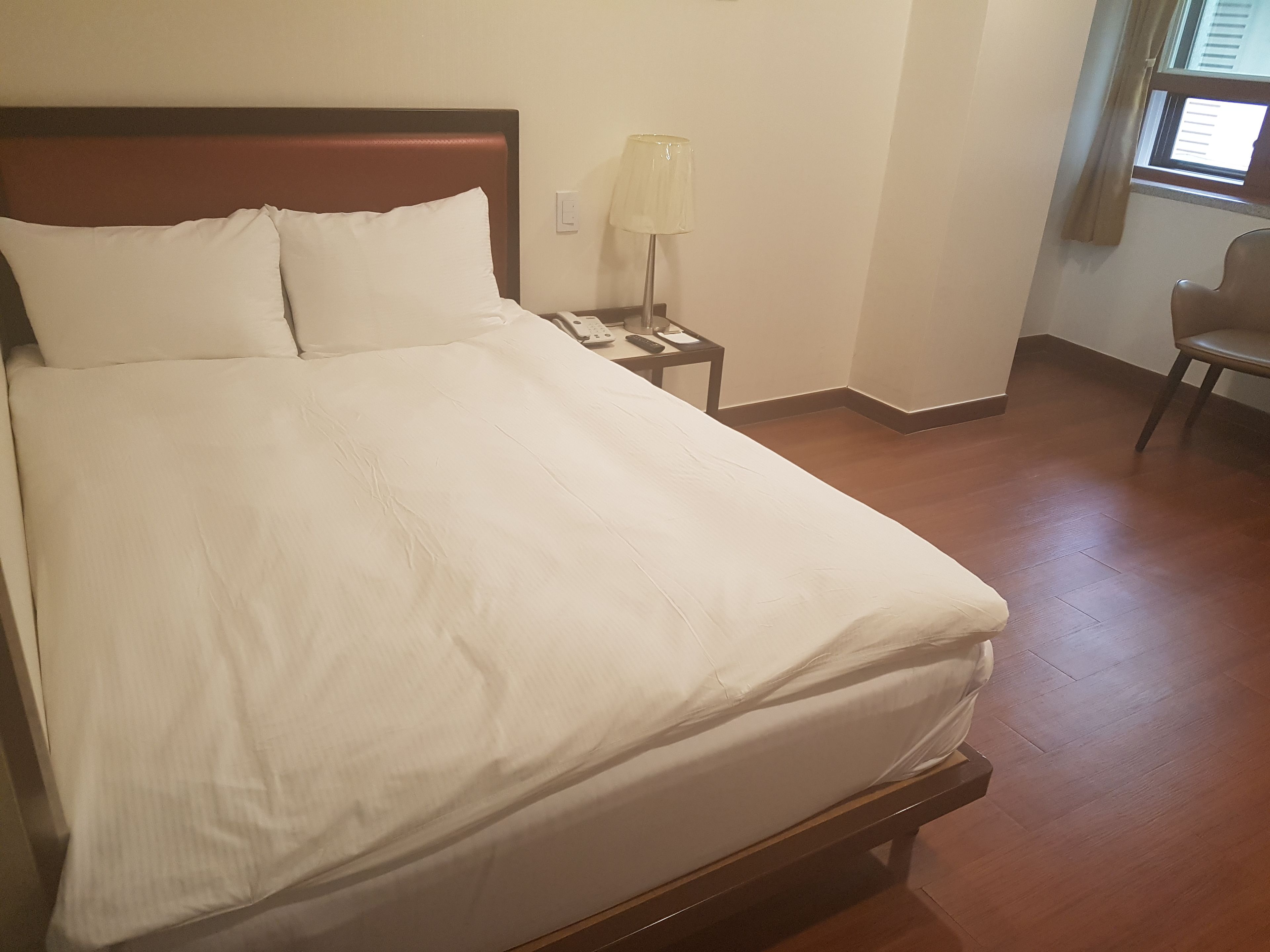 A neatly made bed with white linens, a bedside table with a lamp, and a chair in a room with wooden flooring and a window with blinds.