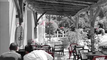 An outdoor café with tables and chairs under a wooden pergola, overlooking a garden with a fountain and trees.