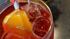 A close-up of a cocktail with ice cubes, a slice of orange, and a decorative orange flower on a striped straw.