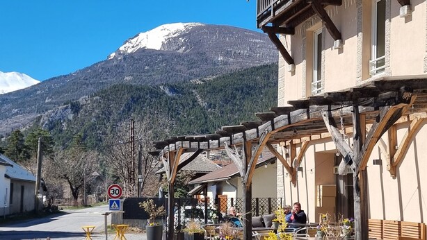 A café with outdoor seating, a sign reading 'Pause de Cycliste,' and a mountain in the background.