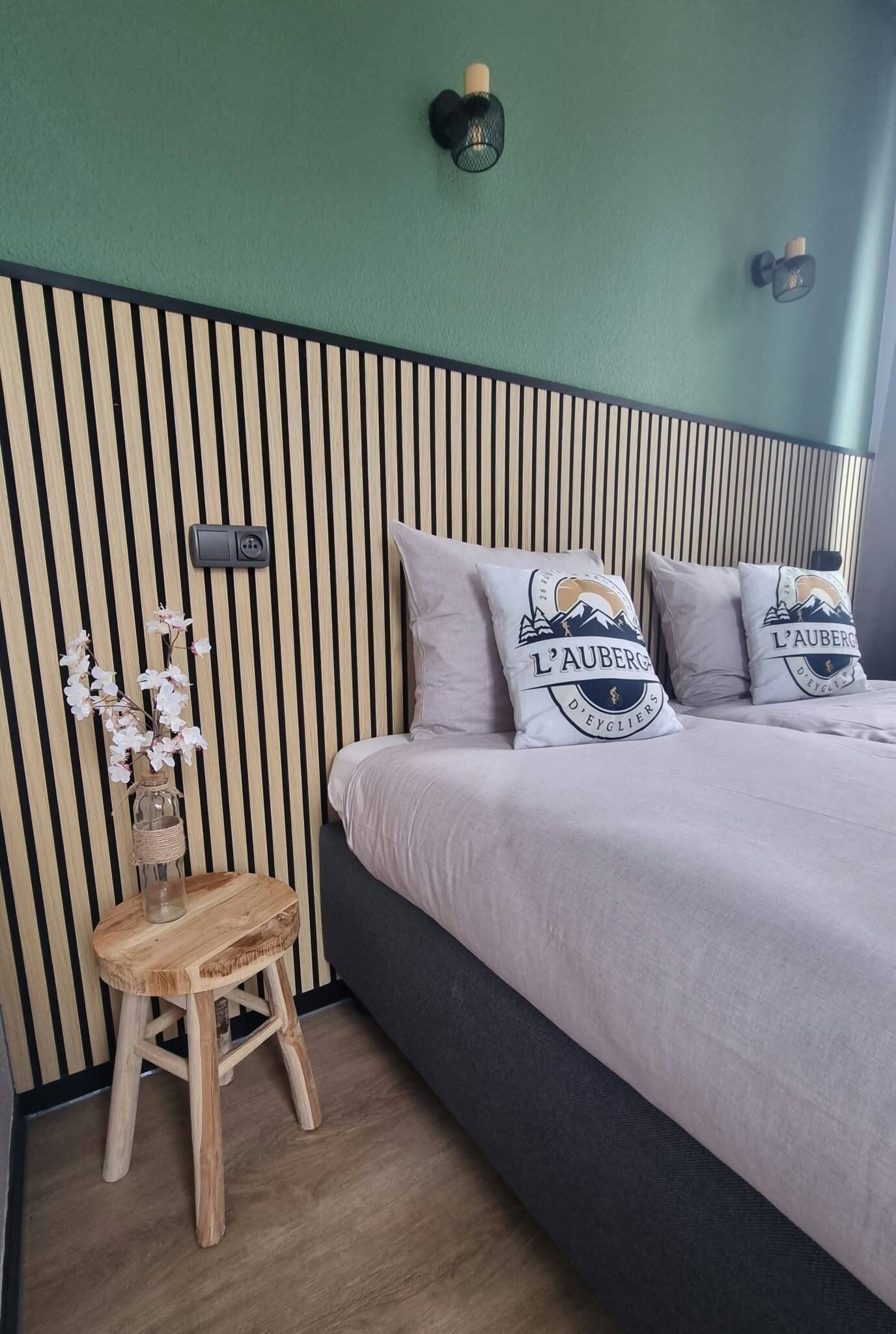 Un dormitorio con cama, almohadas, una mesita de noche, un florero con flores y una lámpara de pared.