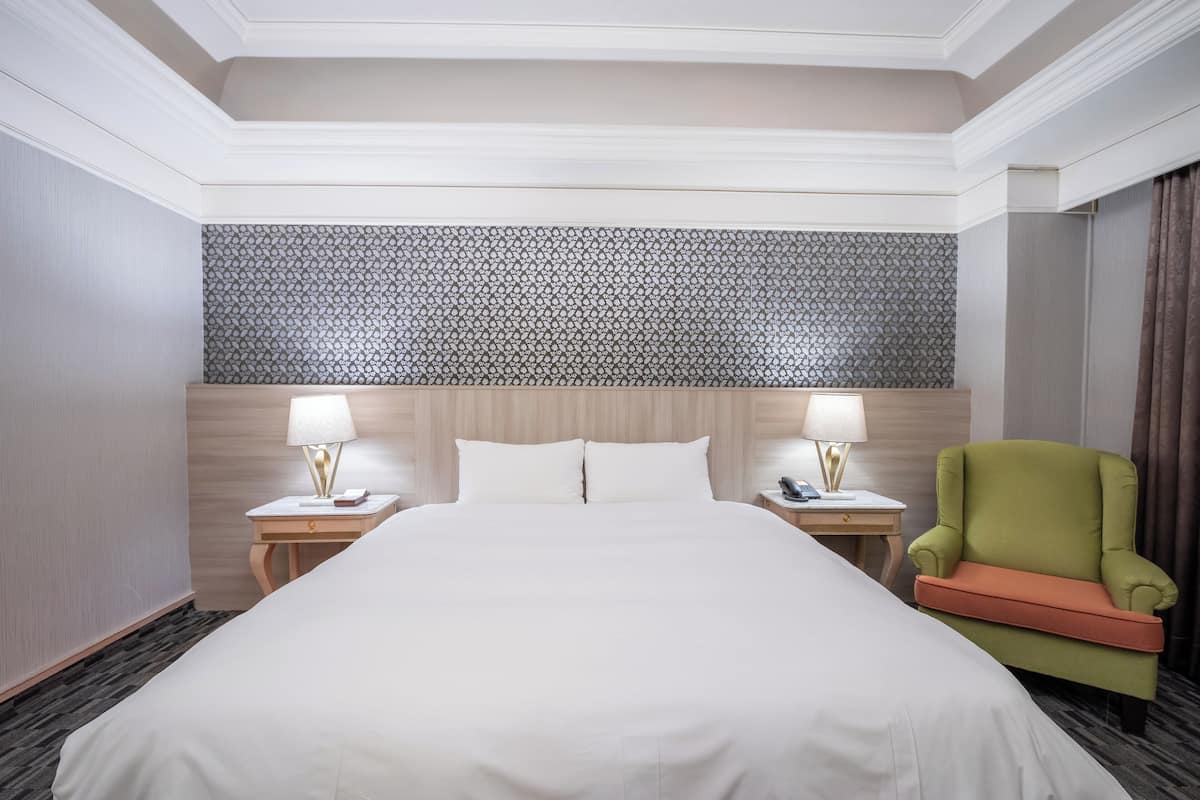 A hotel room with a large bed, two bedside tables with lamps, a green armchair, and a patterned headboard.