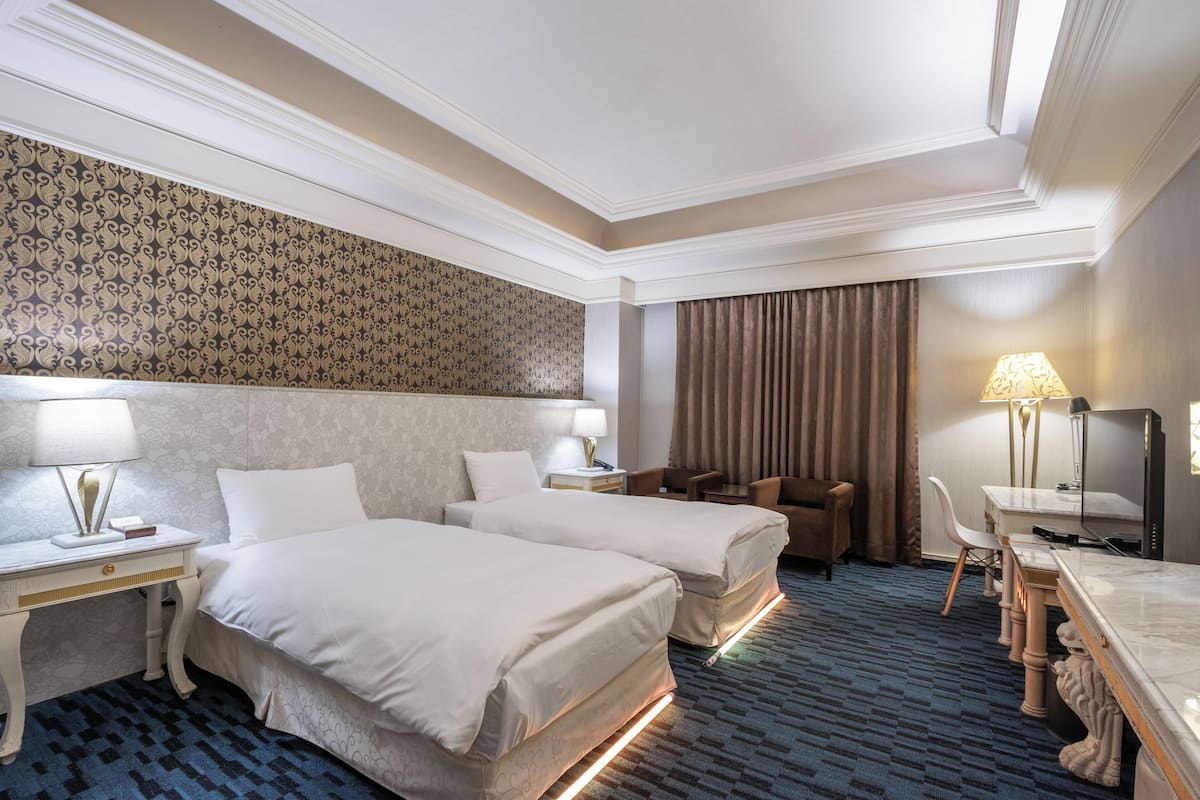 A hotel room with two beds, a desk, a chair, a TV, and a lamp.