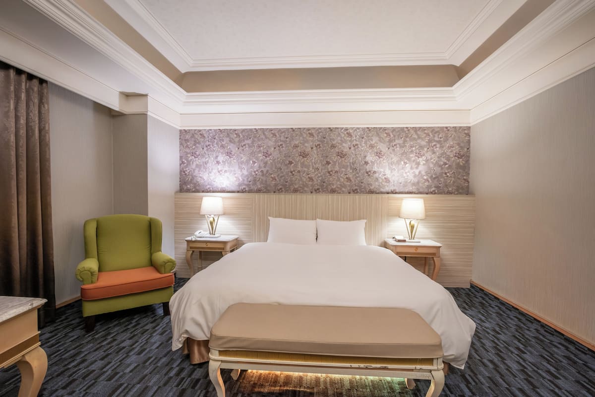 A hotel room with a large bed, two bedside tables with lamps, a green armchair, and a patterned wallpaper headboard.