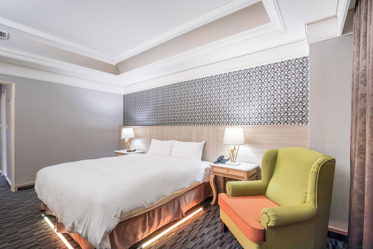A hotel room with a large bed, bedside tables with lamps, a green armchair, and a patterned wall.
