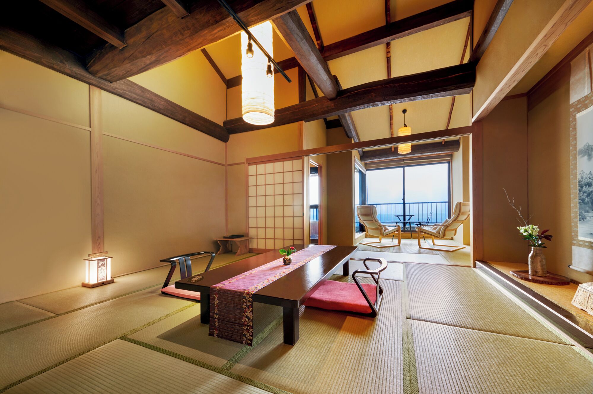A traditional Japanese room with tatami flooring, a low table with a red mat, and a view of a balcony with chairs.