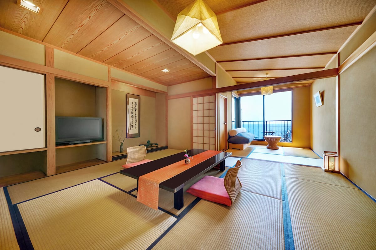 Ett traditionellt japanskt rum med tatamimattor på golvet, ett lågt bord, kuddar och en skjutdörr som leder ut till en balkong med utsikt.