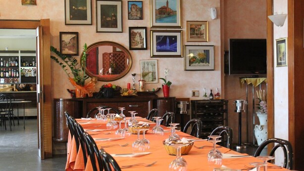 A dining area with a long table set for a meal, surrounded by chairs. There are framed pictures on the walls, a television, and a bar area with bottles visible in the background.