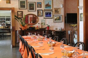 Comedor con una larga mesa dispuesta para una comida, rodeada de sillas. Hay cuadros en las paredes, un televisor y una zona de bar con botellas visibles al fondo.