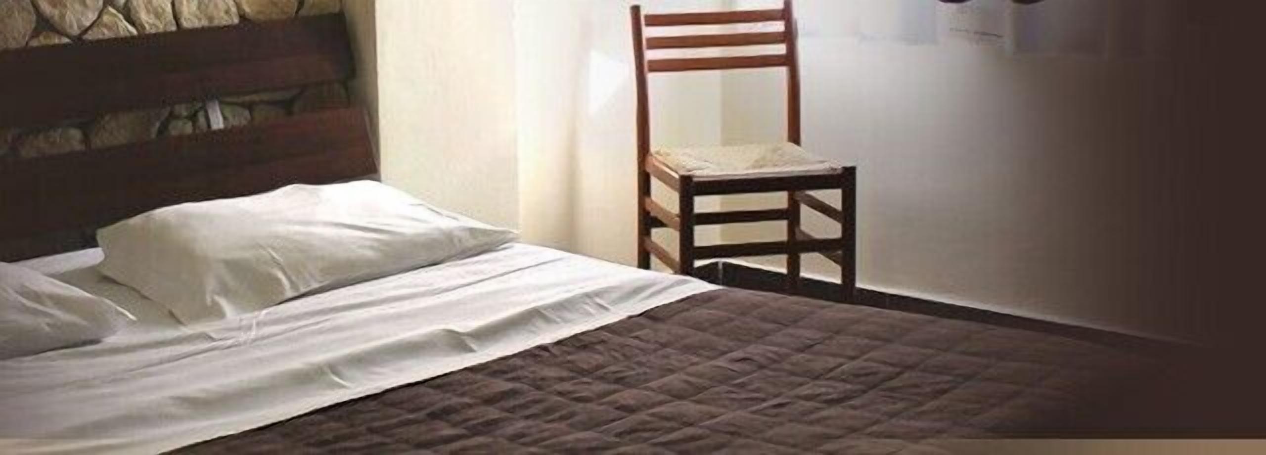 A bed with white linens and a brown quilt, a wooden chair, and a stone wall.