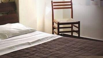 A bed with white linens and a brown quilt, a wooden chair, and a stone wall.