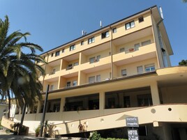 Un hotel de varios pisos con balcones, un cartel que indica que ofrece bar y restaurante, y un anuncio para una trattoría y una tienda de frutas.