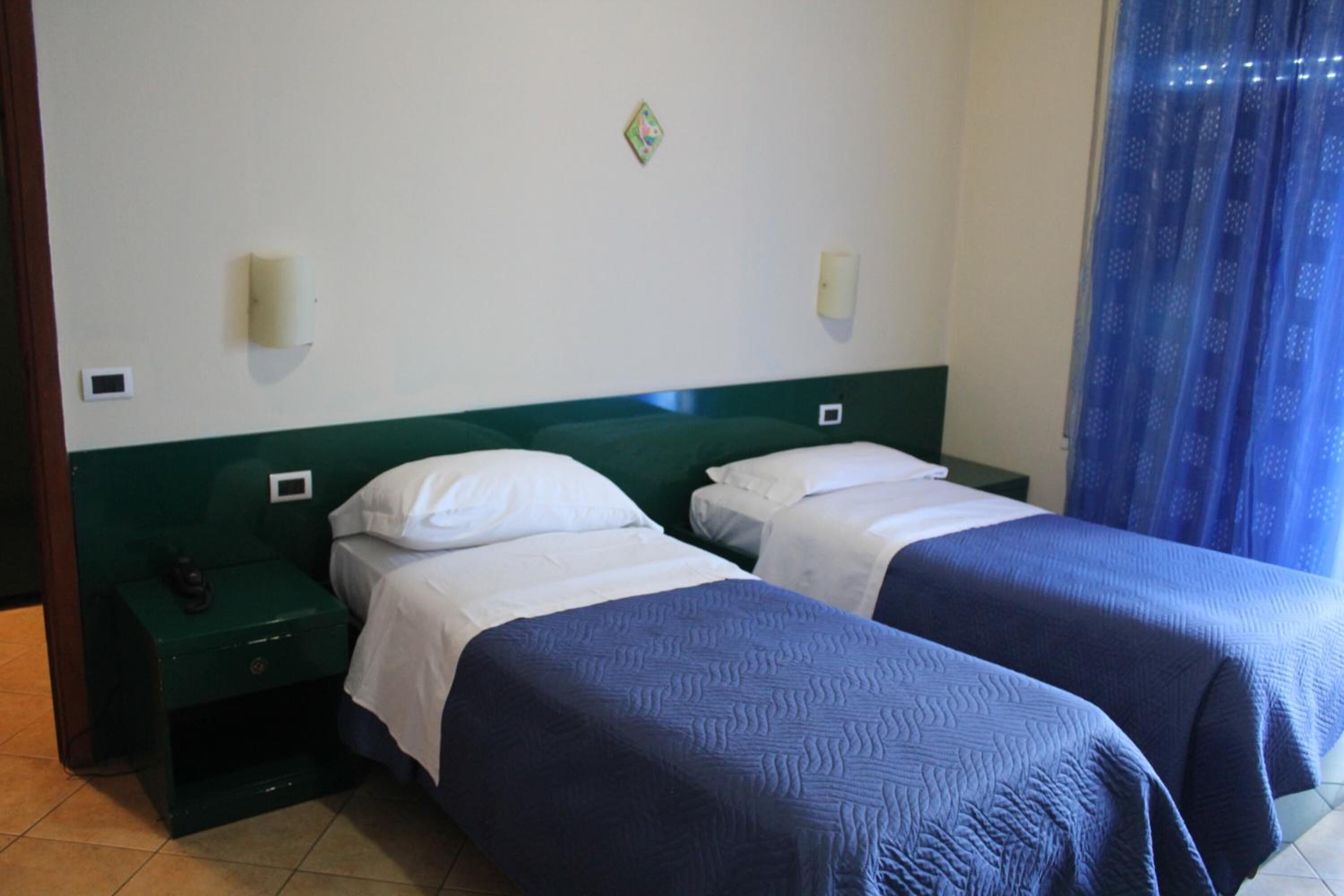 A hotel room with two single beds, a green headboard, white bedding, a nightstand with a phone, and a small decorative item on the wall.