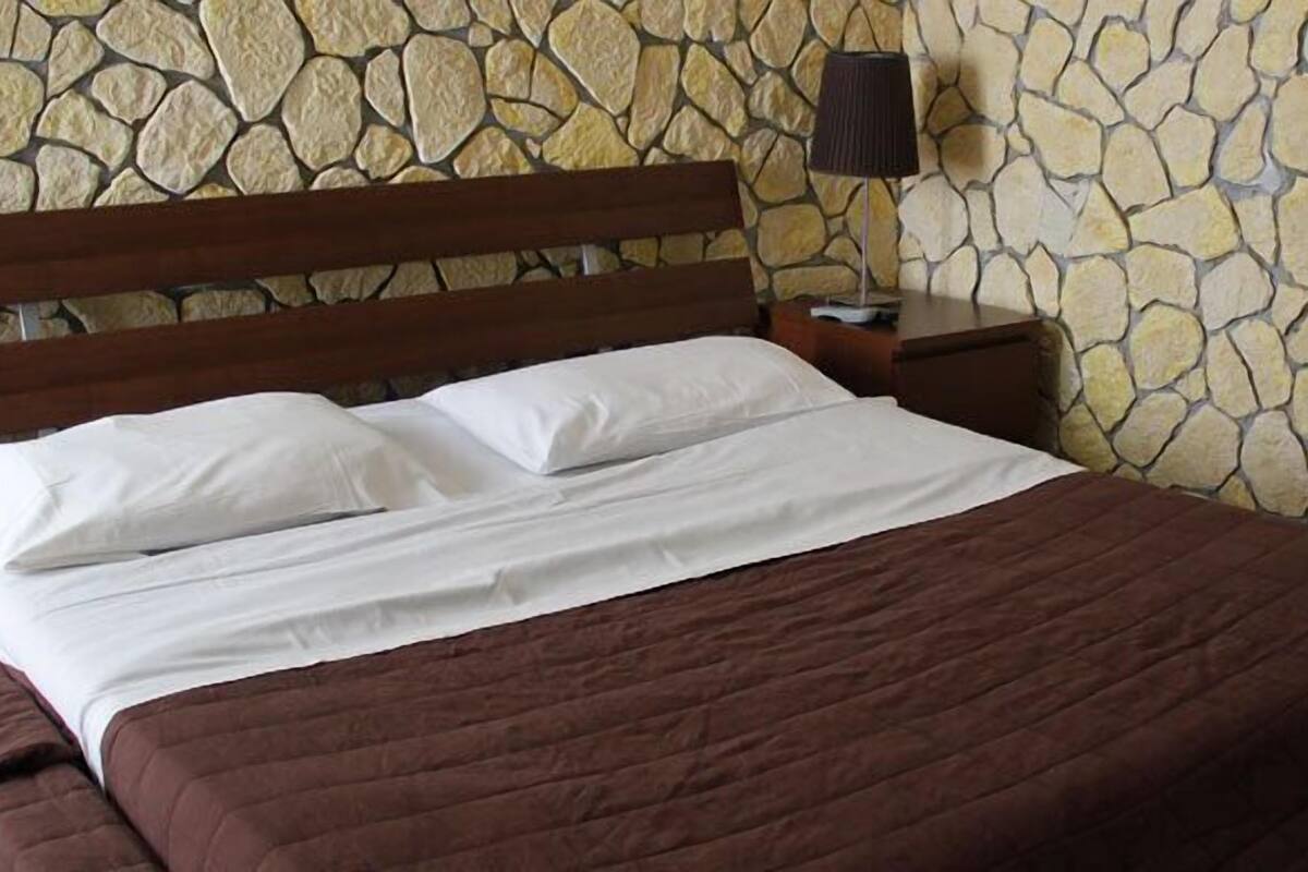 A bed with a wooden headboard, white pillows, and a brown bedspread in a room with a stone wall.